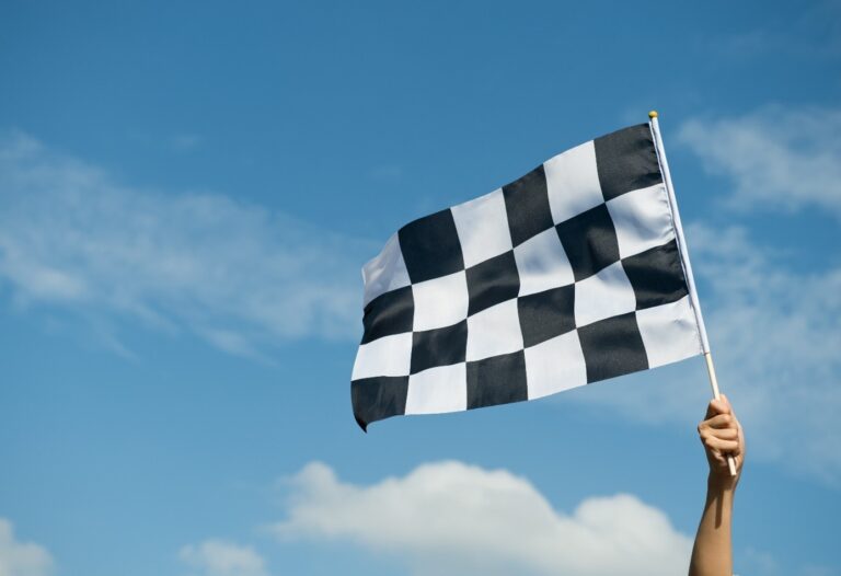 A black and white checkered flag is being held up against a clear blue sky with a few scattered clouds.