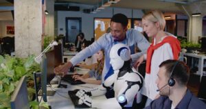 A diverse group of colleagues gather around a humanoid robot working at a computer in a modern office. One man points at the screen while others, including a woman and another man wearing a headset, look on attentively. Green plants decorate the spacious office.