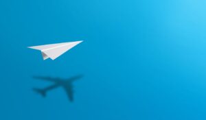 Un avión de papel volando alto en el cielo con su sombra en forma de avión real debajo, sobre un fondo azul claro.