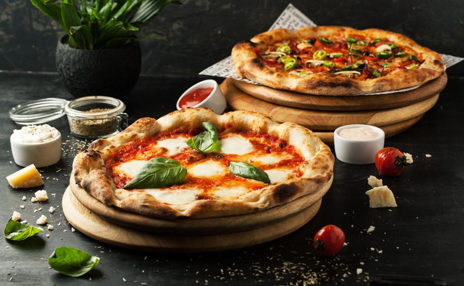 Two fresh pizzas on wooden boards, one with basil and cheese, and the other with various toppings, surrounded by ingredients.