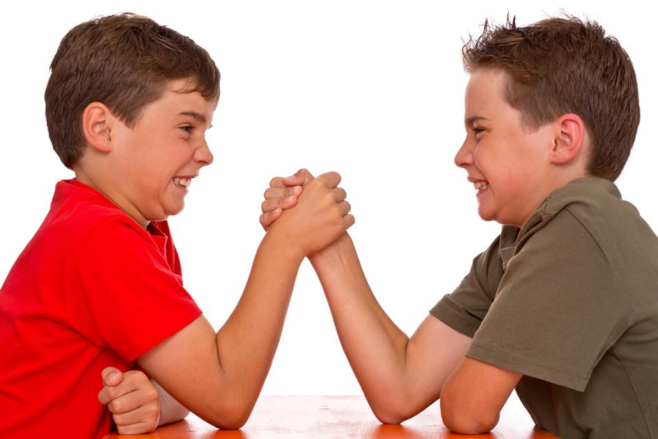 Two young boys are engaged in an arm-wrestling match, smiling with determination. One is wearing a red shirt, and the other is wearing a green shirt. They are seated at a table, clasping hands tightly.