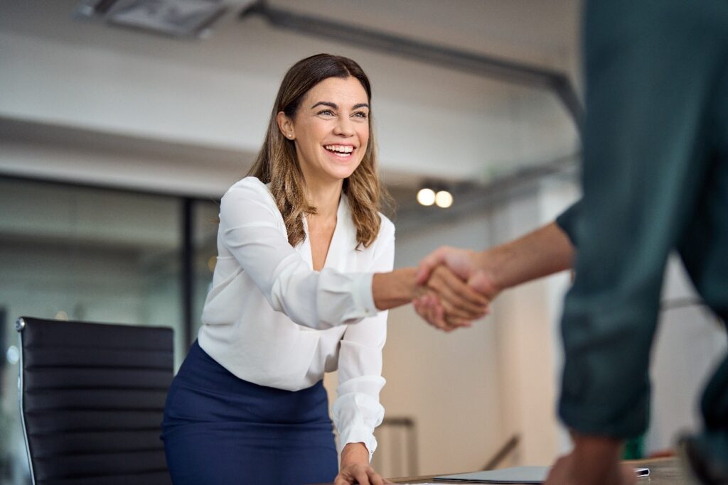 A professional woman wearing a white blouse and blue skirt is smiling warmly and shaking hands with someone, likely in a business meeting or interview setting. The background shows an office environment with glass walls and modern furniture.