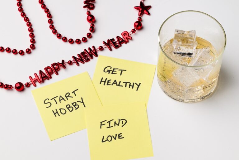 The image shows a festive "Happy New Year" decoration in red, a glass of whiskey with ice, and three yellow sticky notes with New Year resolutions written on them: "Start Hobby," "Get Healthy," and "Find Love."