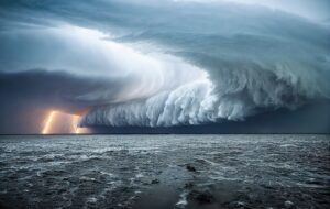 A massive storm cloud with a dramatic, wave-like formation looms over the ocean. Bright lightning bolts illuminate the dark sky, casting an eerie glow on the turbulent water below.