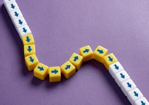 A chain of small white and yellow cubes with blue arrows is arranged on a purple background. The white cubes form a straight path, while the yellow cubes introduce a curved deviation, symbolizing a disruption or change in direction.