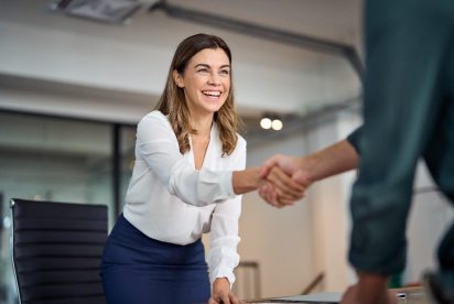 A professional woman wearing a white blouse and blue skirt is smiling warmly and shaking hands with someone, likely in a business meeting or interview setting. The background shows an office environment with glass walls and modern furniture.