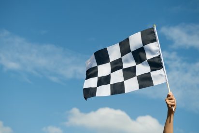 Une main tient un drapeau à damier noir et blanc sur un ciel bleu avec quelques nuages. Le drapeau, souvent associé aux courses automobiles, flotte dans les airs.