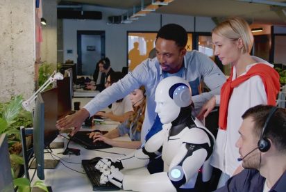 Dans un bureau moderne, un groupe diversifié de personnes collabore avec un robot humanoïde. Une personne montre un écran d’ordinateur pendant que d’autres observent. Le décor regorge d’activités, de plantes et d’équipements de bureau en arrière-plan.
