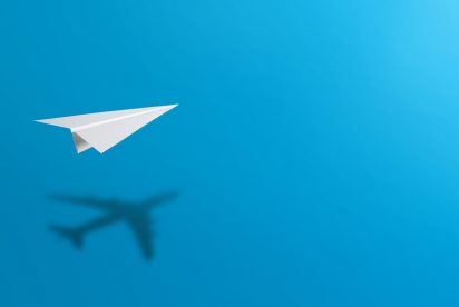 Un avion en papier au premier plan avec son ombre ressemblant à un véritable avion projetée sur un fond bleu vif.