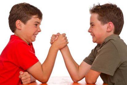 Two young boys are engaged in an arm-wrestling match, smiling with determination. One is wearing a red shirt, and the other is wearing a green shirt. They are seated at a table, clasping hands tightly.