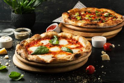 Two fresh pizzas on wooden boards, one with basil and cheese, and the other with various toppings, surrounded by ingredients.